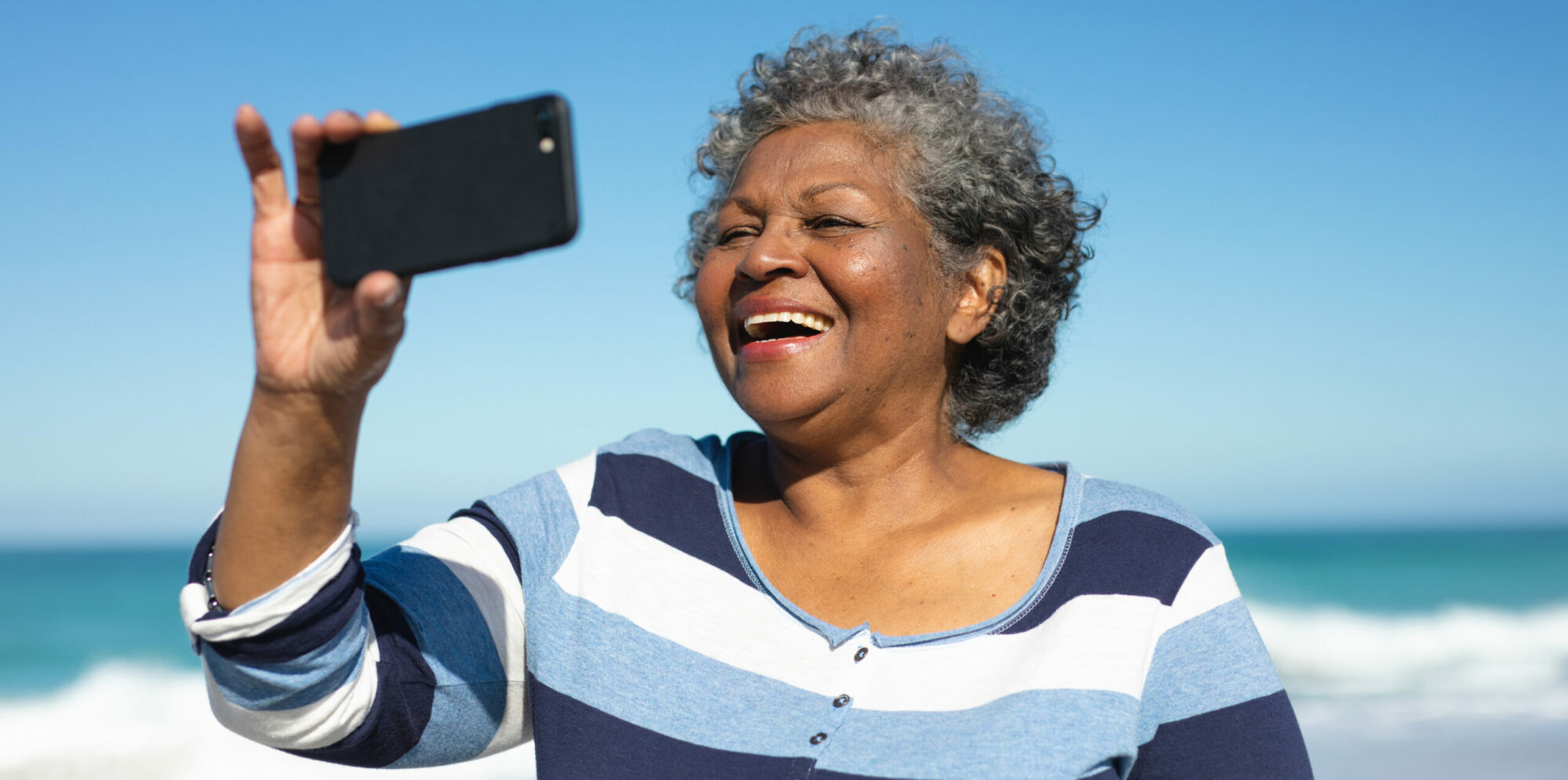 Front view close up of a senior African American woman standing on the beach with blue sky in the background, laughing and taking a selfie with a smartphone